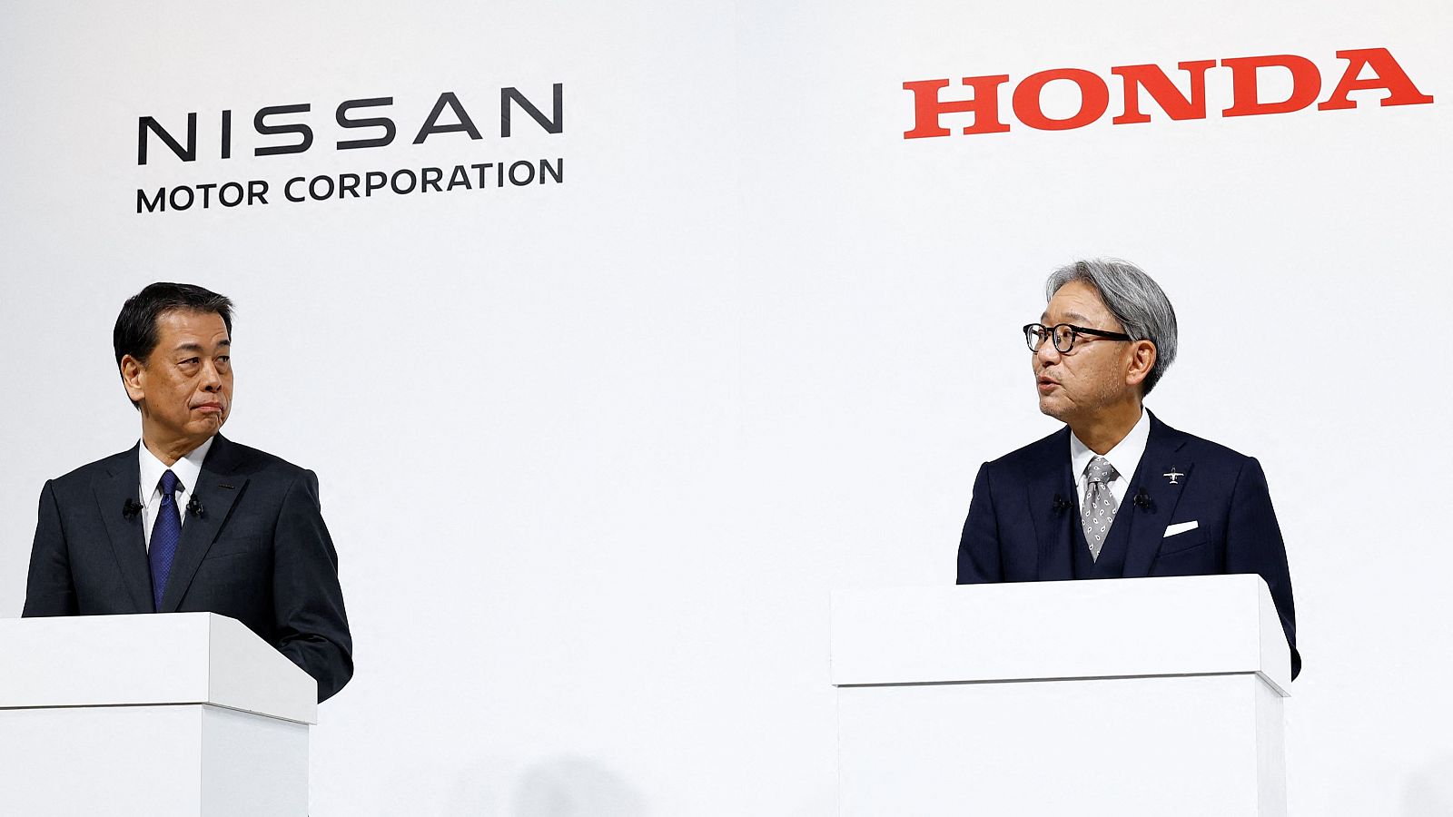 Conferencia de prensa o evento similar con dos hombres en traje detrás de podios blancos.  Logotipos de Nissan y Honda visibles en el fondo.