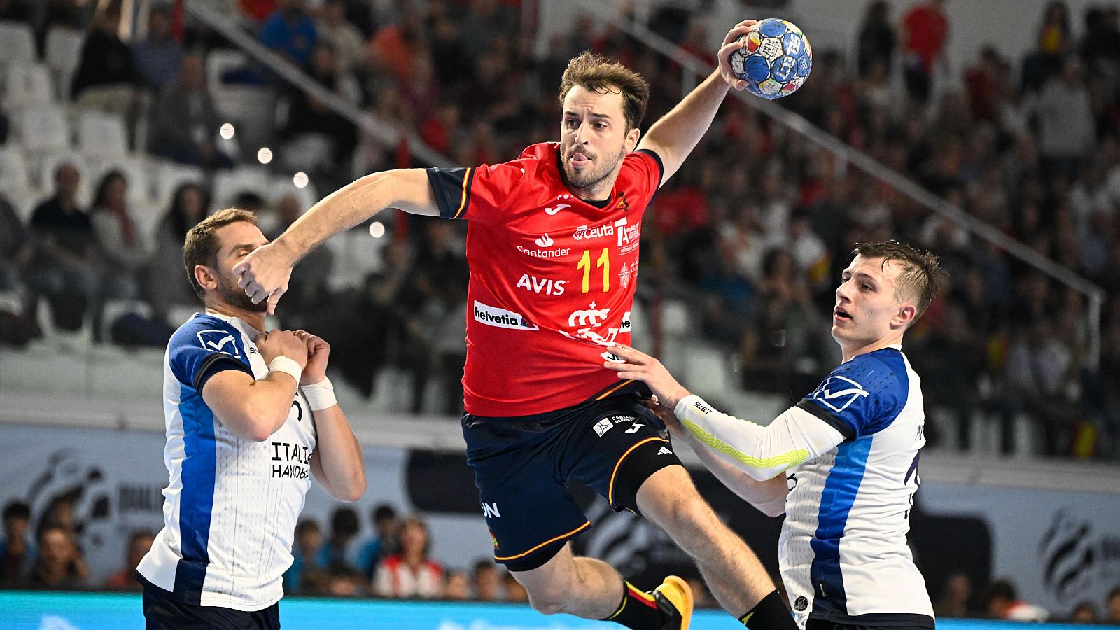 Jugador de balonmano español (camiseta roja, dorsal 11) salta para lanzar, esquivando a dos rivales (camisetas blancas y azules).  Posiblemente, partido del Torneo Internacional.