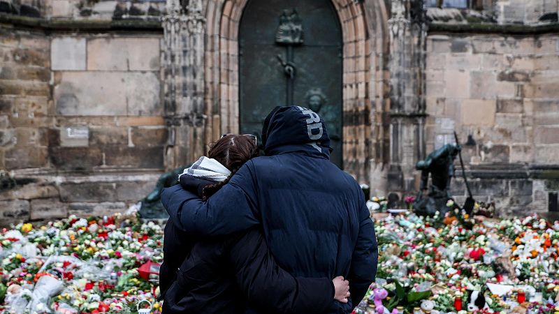 Pareja abrazada ante extensa ofrenda floral con flores, velas y peluches, frente a un edificio de piedra.  Una persona lleva una chaqueta con la palabra "HARDY".