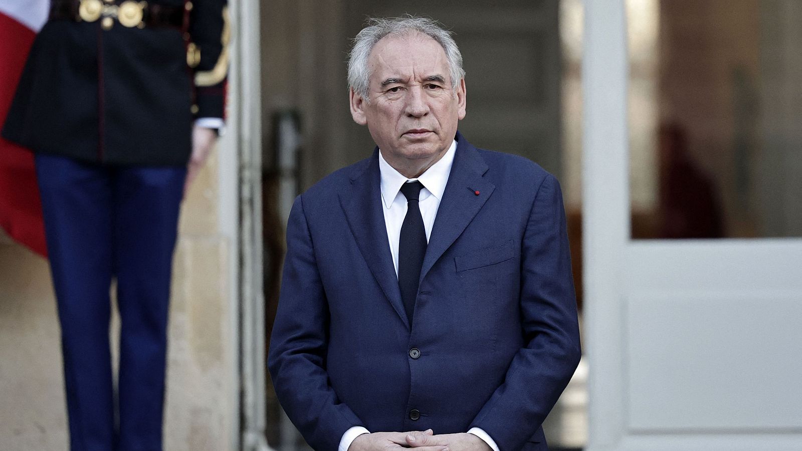 Imagen de François Bayrou en traje azul marino, camisa blanca y corbata negra, con una persona uniformada y bandera francesa al fondo. Posible anuncio político.