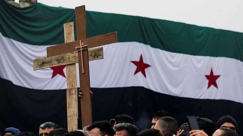 Cruz de madera en manifestación con bandera siria al fondo. Protesta por la quema de un árbol de Navidad, mostrando defensa de la libertad religiosa.