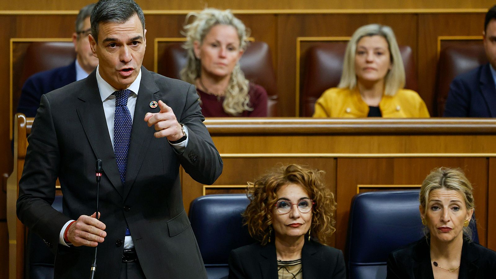 El presidente del Gobierno español, de traje oscuro y corbata, interviene en el Congreso, gesticulando mientras habla. Dos mujeres observan desde la parte inferior.