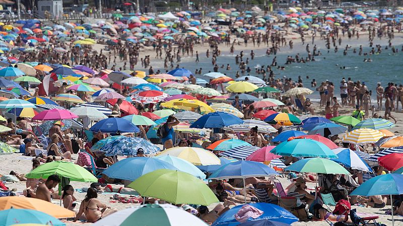 Playa llena de gente y sombrillas de colores.  Se observa el mar al fondo y algunas estructuras. Alta afluencia turística.