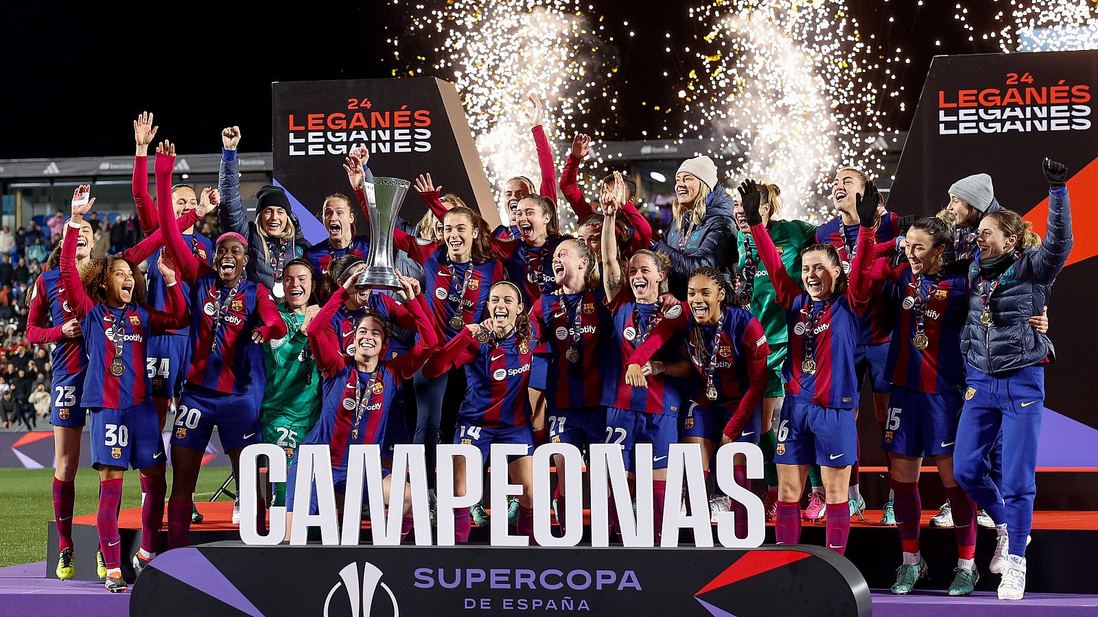 Celebración del equipo femenino de fútbol tras ganar un torneo. Jugadoras con medallas y trofeo, confeti y fuegos artificiales de fondo.  Se lee "CAMPEONAS" en letras grandes.