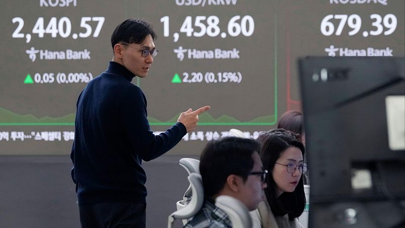 Operador de divisas señalando pantalla con datos financieros: 2,440.57 (0.00%), 1,458.60 (0.15%), 679.39.  Ambiente de trabajo en sala de operaciones.