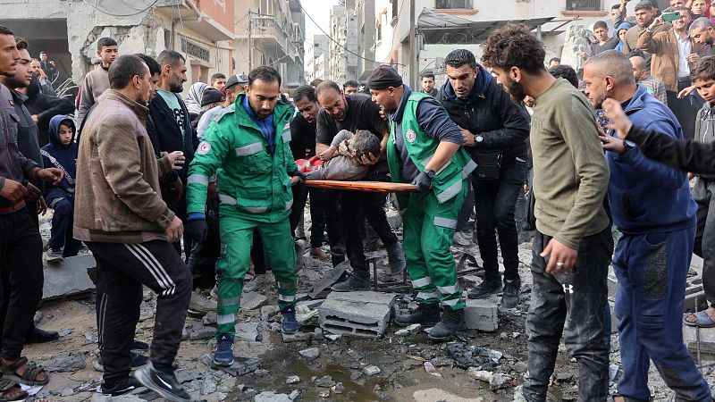Paramédicos trasladan a un herido en camilla por una calle con escombros.  Multitud observa.  Escena de urgencia y caos.