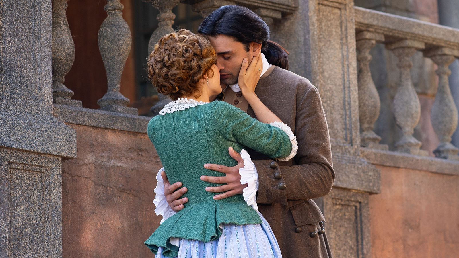 Pareja abrazada y besándose apasionadamente en un entorno exterior con balaustrada.  Ella viste un traje de época verde oscuro y azul, él una chaqueta marrón.