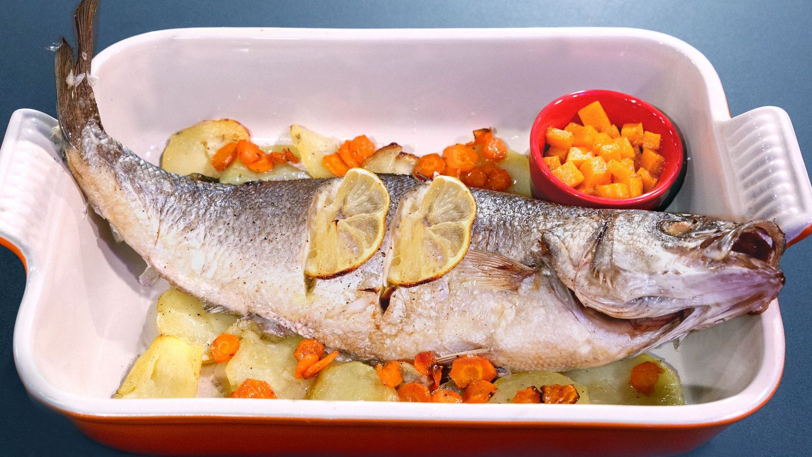 Lubina dorada al horno con patatas, zanahorias y dados de calabaza.  Presentada en fuente blanca con limón.