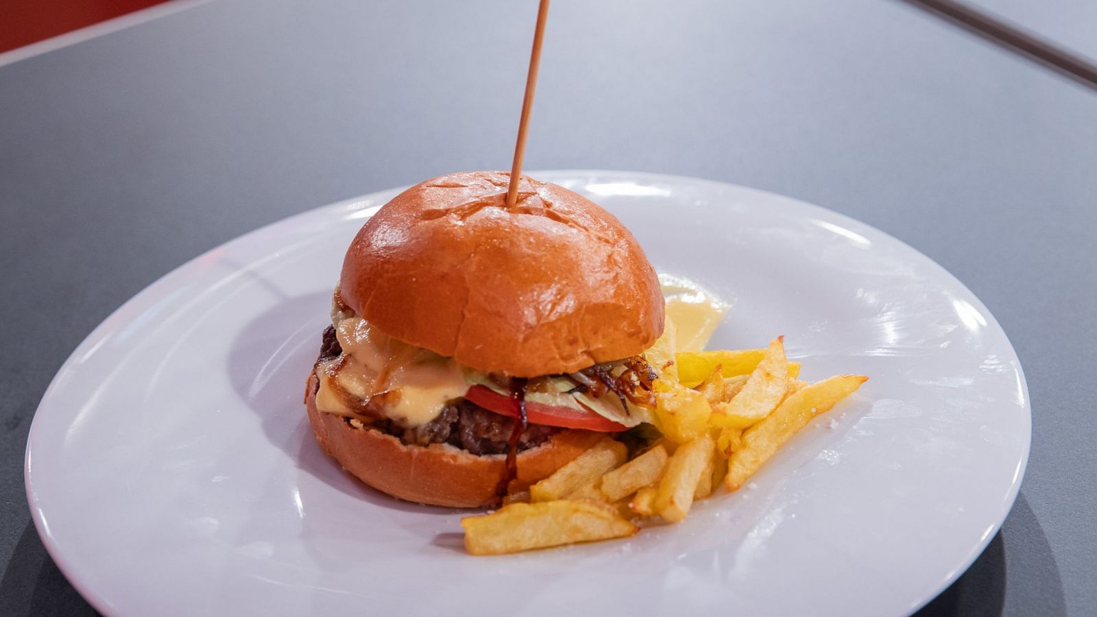 Hamburguesa con pan brioche, carne, queso, tomate, cebolla y salsa, acompañada de patatas fritas en un plato blanco.