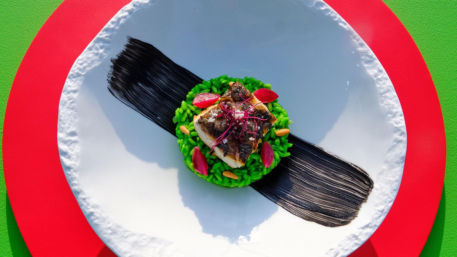 Corvina soasada con arroz verde, alioli negro y detalles rojos sobre plato blanco.  Presentación culinaria sofisticada y colorida.