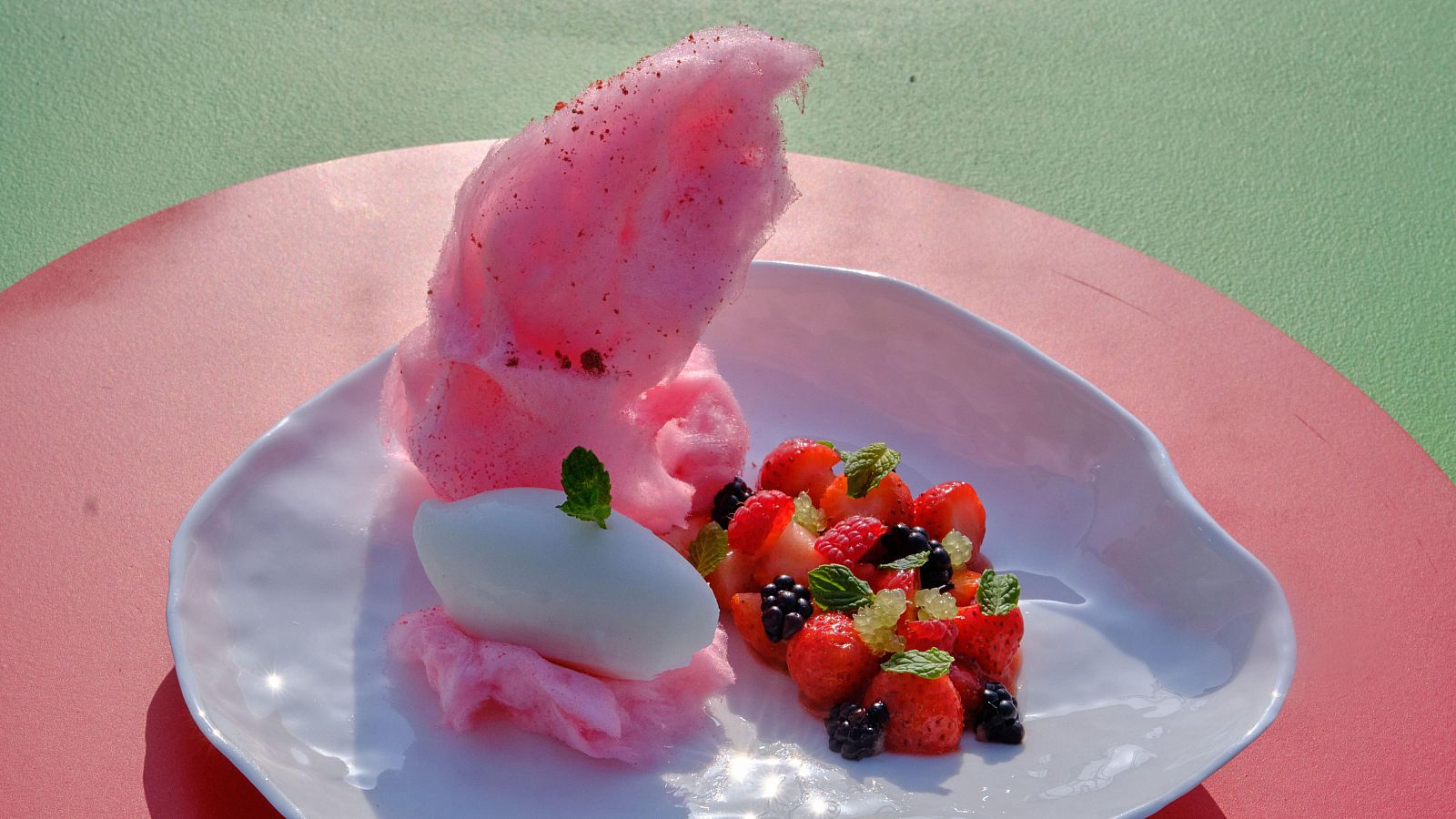 Postre de MasterChef Junior con frutos rojos, caviar cítrico y algodón de azúcar rosa sobre una base blanca.  Menta fresca decora el plato.