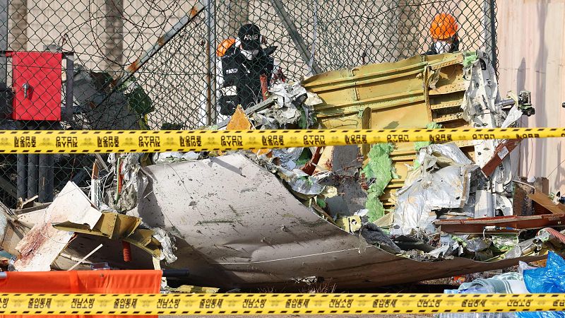 Restos de avión accidentado: fuselaje, alas y otros componentes rotos y dispersos. Personal de emergencia en la escena acordonada.