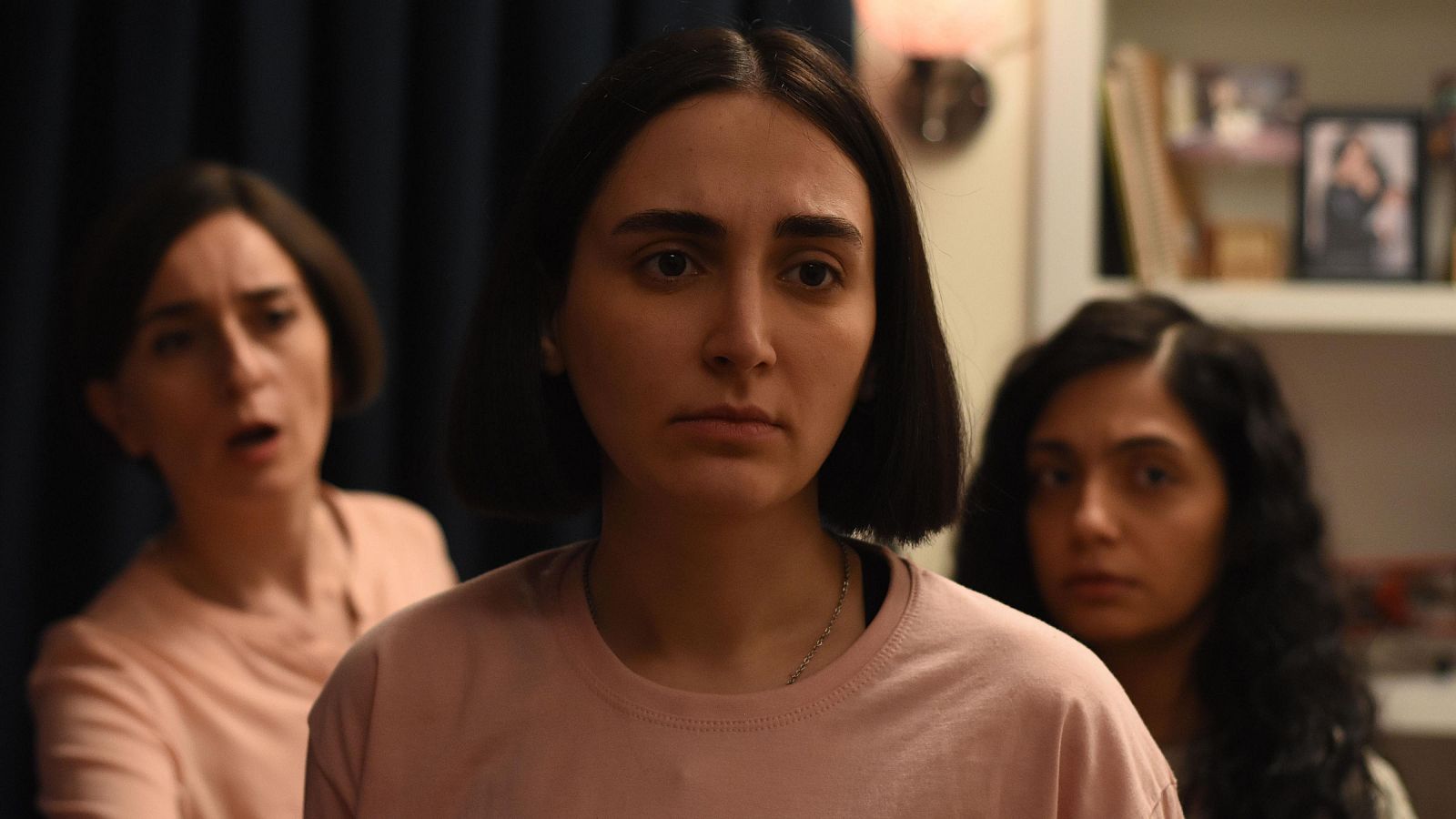 Tres mujeres, una con camiseta rosa en primer plano mirando a cámara, las otras dos detrás, ligeramente desenfocadas.  Ambiente dramático.