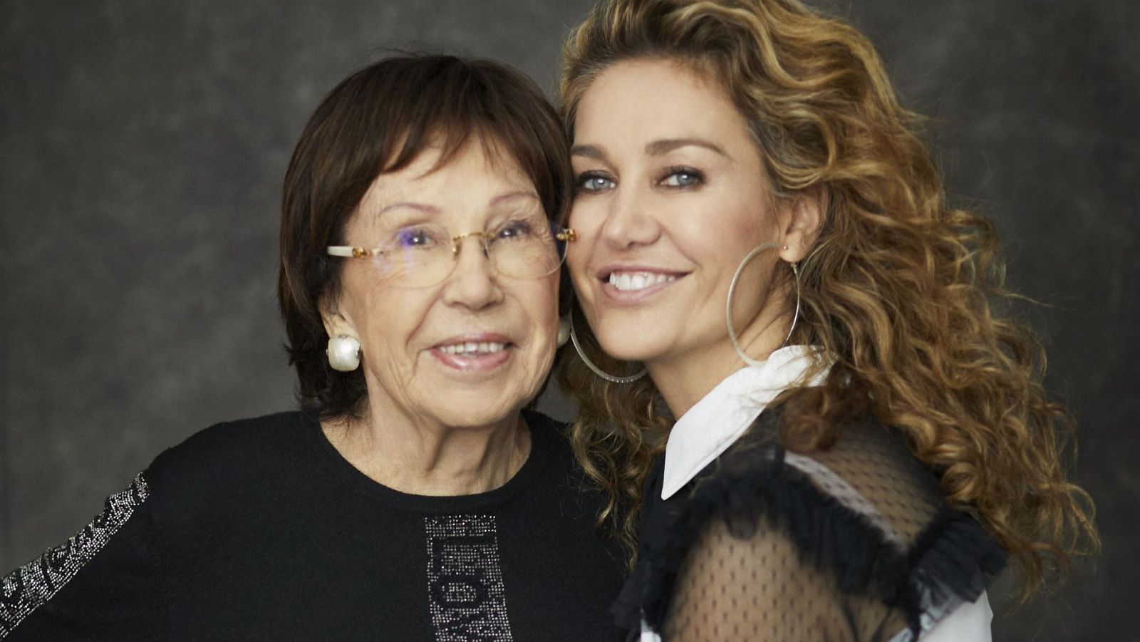 Retrato de dos mujeres sonrientes, madre e hija, con ropa elegante. Una lleva blusa negra y gafas; la otra, blusa blanca con capa negra semitransparente.