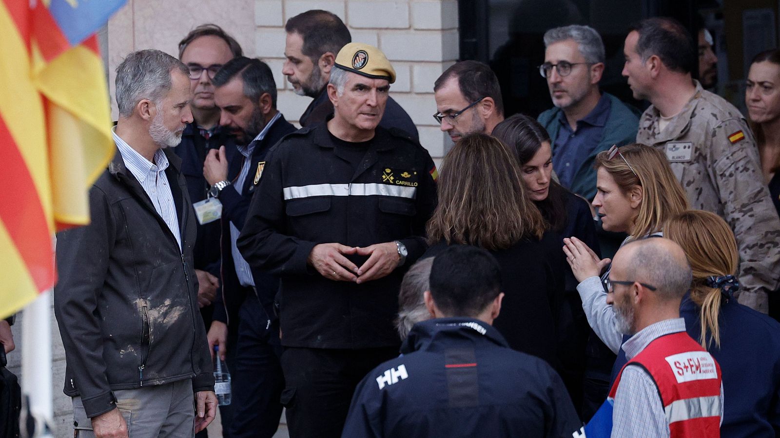 Los Reyes de España visitan un centro de emergencias, donde el Rey viste una chaqueta con barro y la Reina viste de negro.  Autoridades y personal de emergencias les informan.