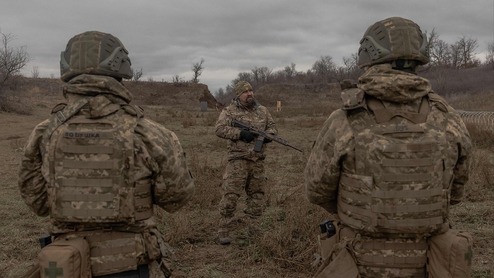 Soldados ucranianos en entrenamiento o zona de combate.  Uniformes de camuflaje, rifles y chalecos antibalas.  Entorno árido con árboles y alambrada.