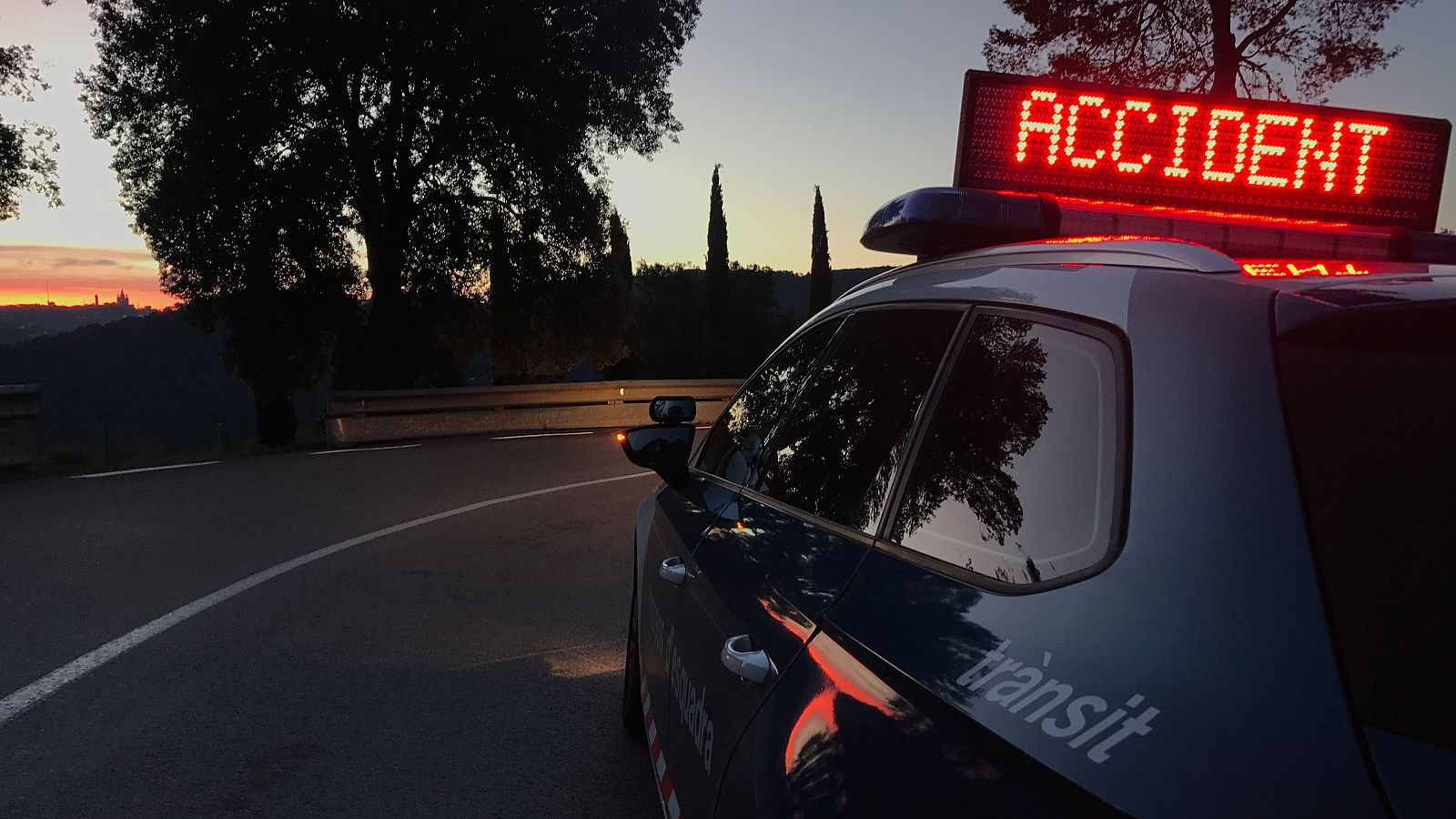 Coche patrulla de los Mossos d'Esquadra señalizando un accidente de tráfico en una carretera con curvas. Panel luminoso con la palabra 'ACCIDENT' visible.