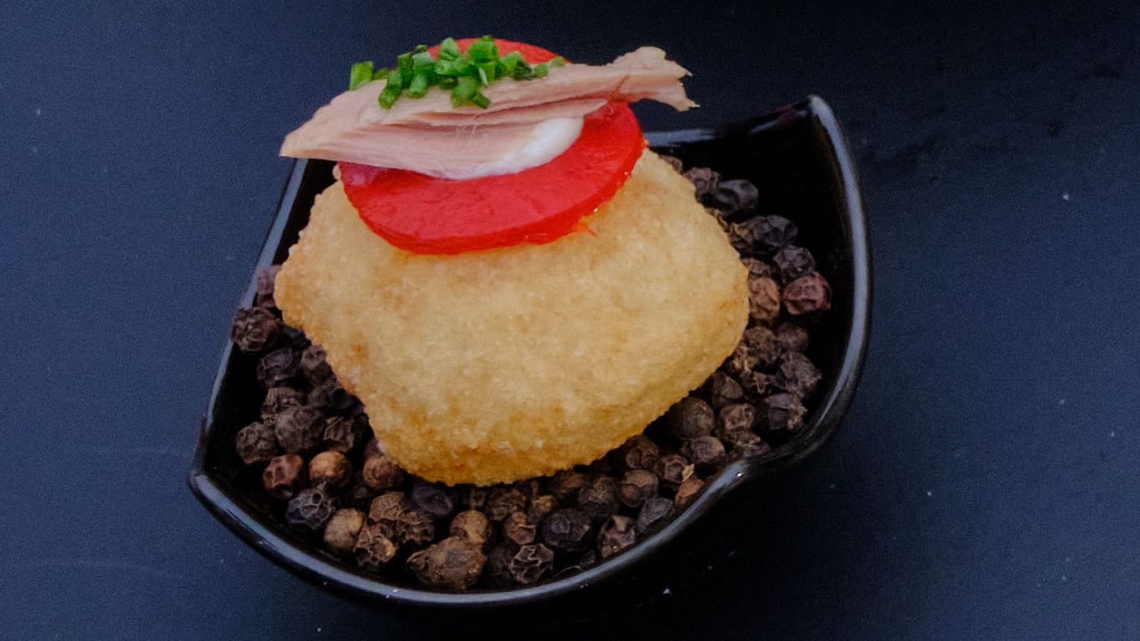 Buñuelo dorado de ensaladilla rusa con tomate, mayonesa, ventresca y cebollino, presentado en un cuenco sobre pimienta negra.