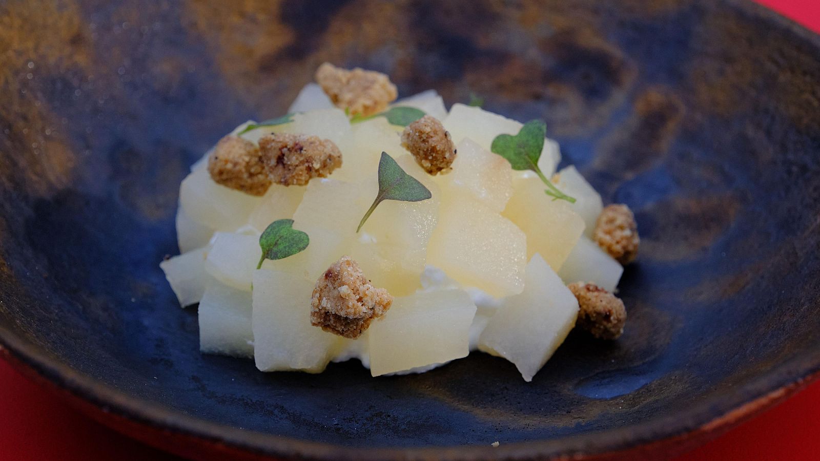 Postre con cubos de pera, crema blanca y crumble marrón en un bol oscuro. Decoración sencilla con hojas verdes.