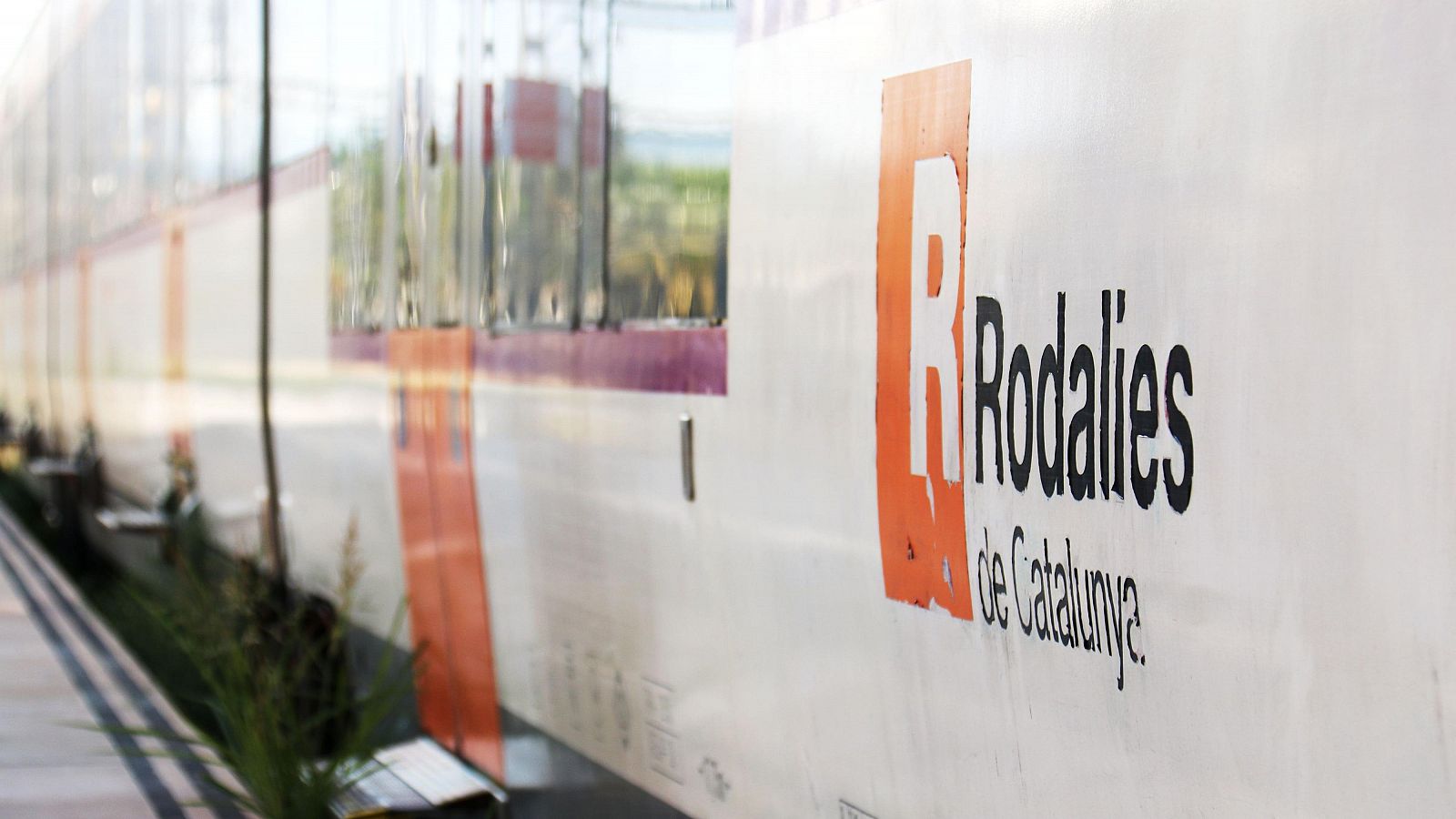 Lateral de un tren Rodalies de Catalunya parado en un andén. Se observa el logo 'R' naranja y el nombre completo. Vegetación desenfocada en primer plano.