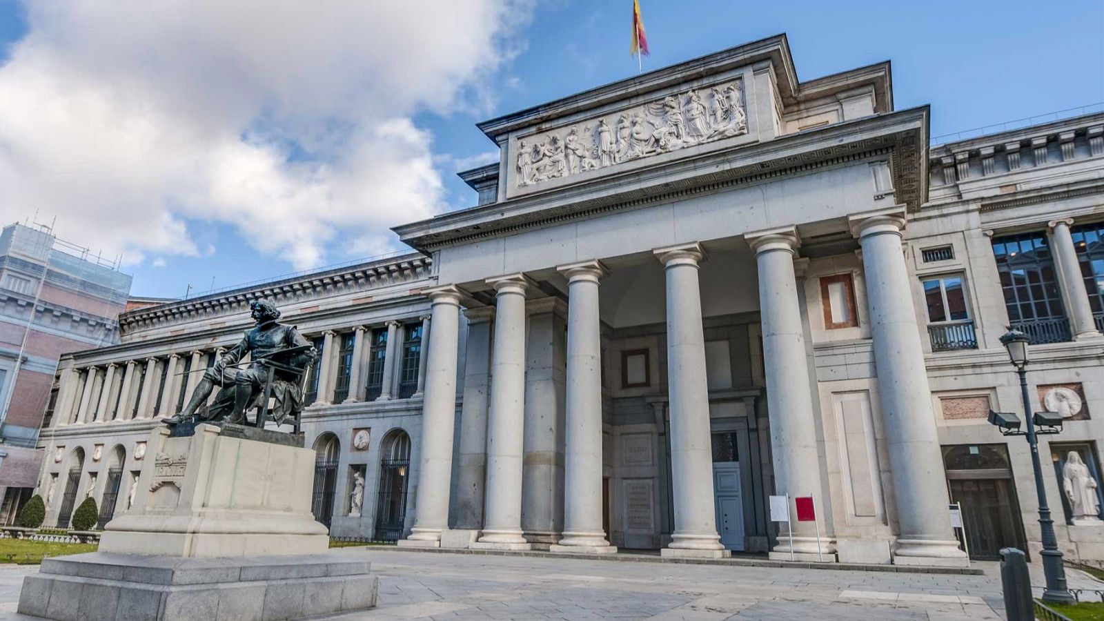 Fachada del Museo del Prado con columnas, frontón y estatua de Goya en primer plano. Bandera española ondeando en el tejado.