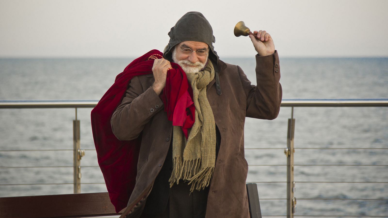 Hombre mayor con barba, gafas y gorro, toca una campana junto al mar. Lleva bufanda y saco rojo.  Escena tranquila y festiva.