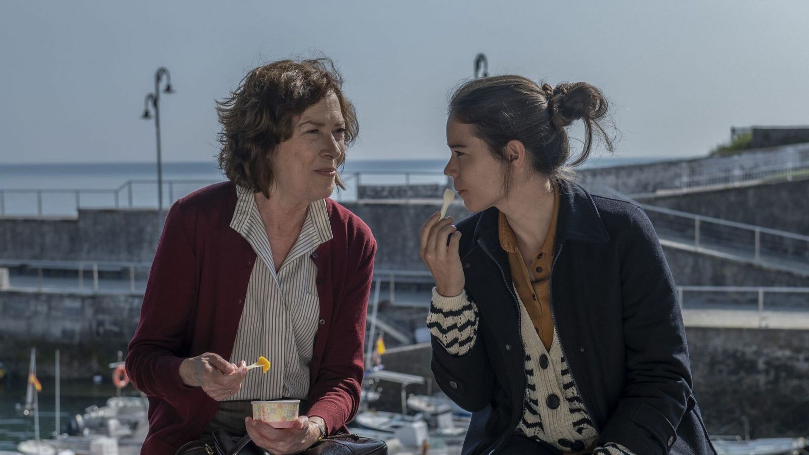 Dos actrices comparten un refrigerio junto al mar, sentadas en un muro. Visten ropa informal y conversan.  Al fondo, barcos y una estructura de hormigón.  Imagen promocional 'Somos cine'.