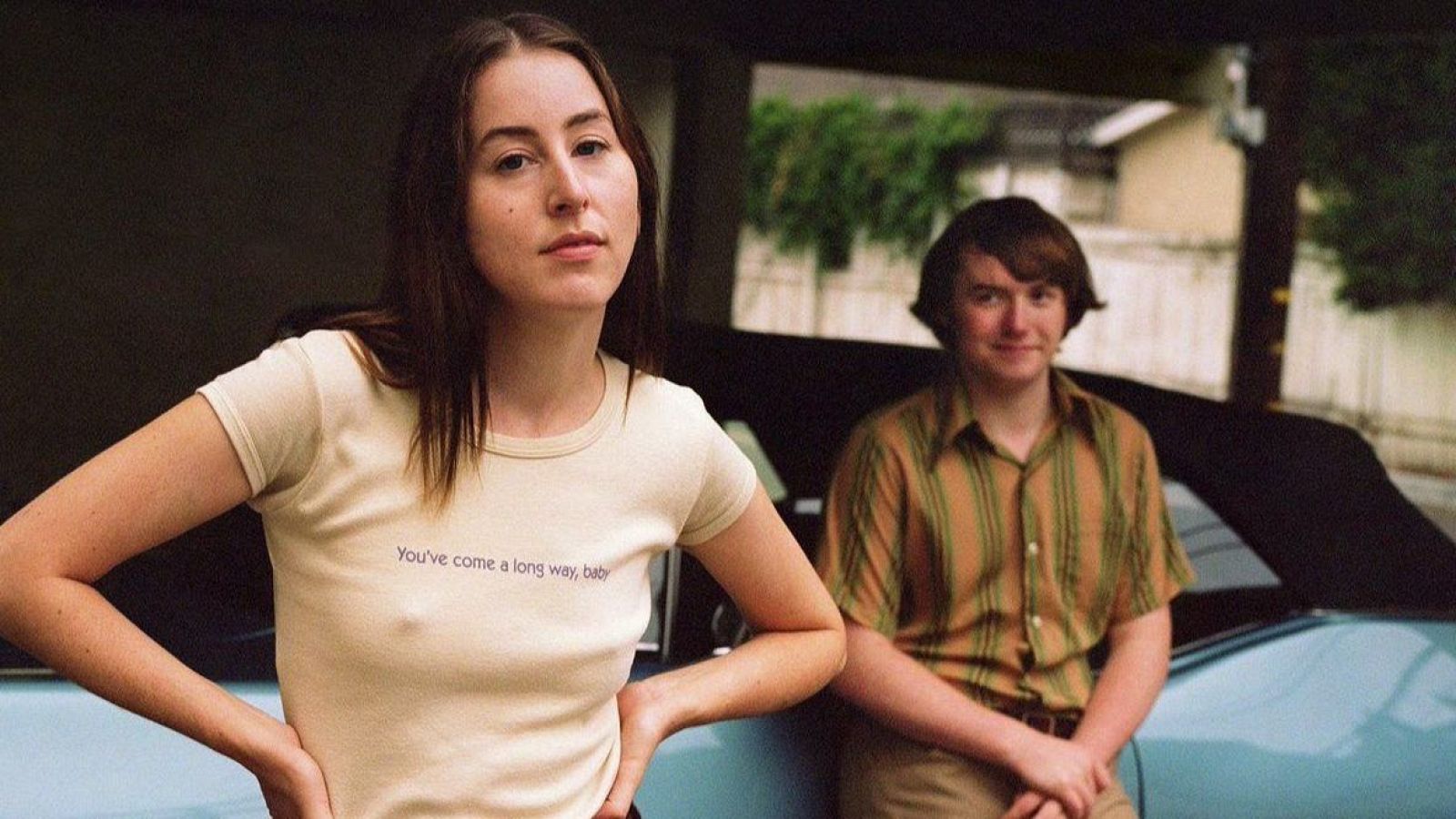 Imagen de Alana Haim y Cooper Hoffman en una escena de *Licorice Pizza*. Ella viste camiseta beige y él camisa a rayas; ambos posan junto a un coche descapotable.