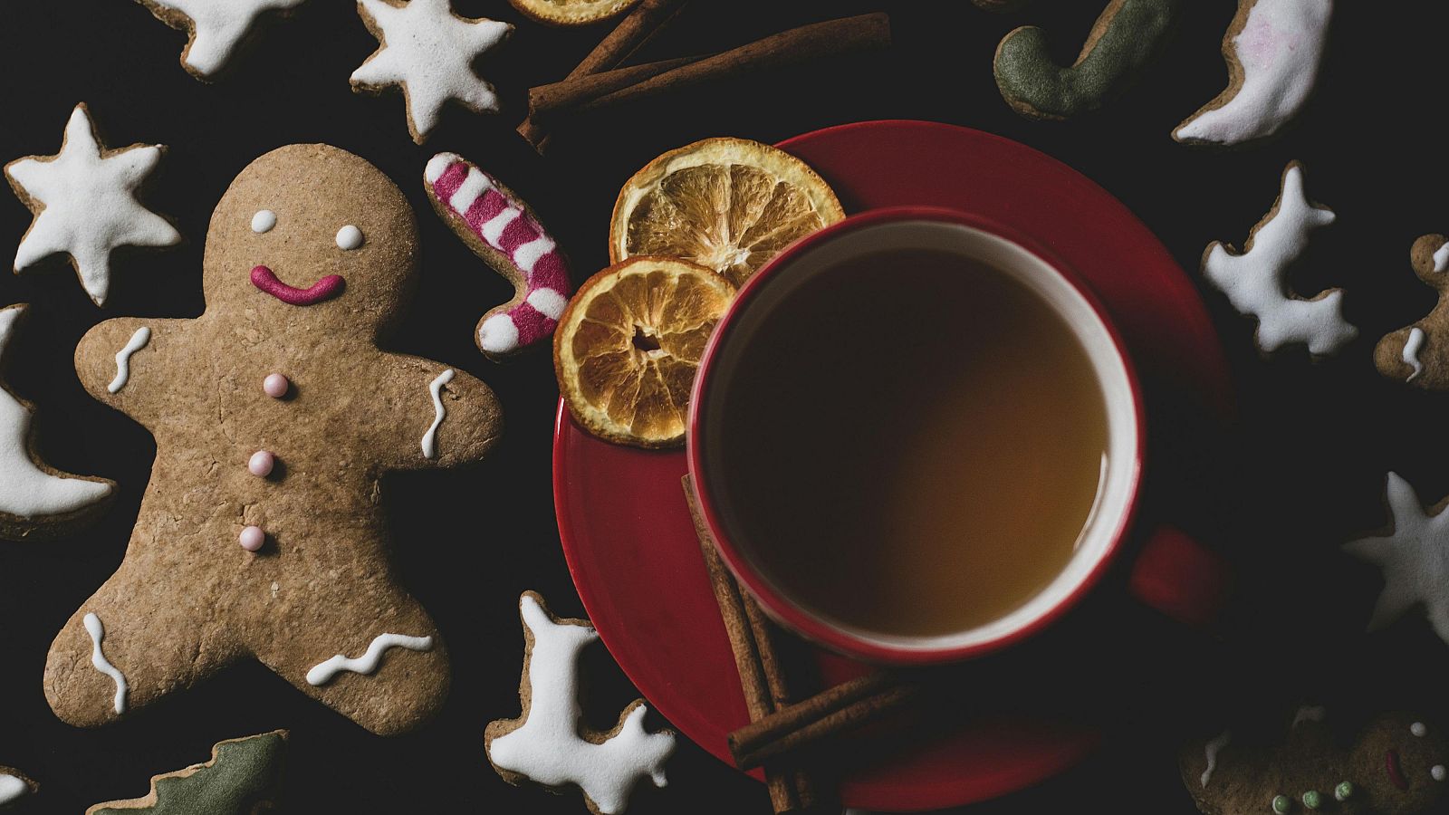 Galletas de jengibre navideñas, hombre de jengibre decorado, bastón de caramelo y otras figuras, junto a una taza roja de té y especias sobre fondo oscuro.