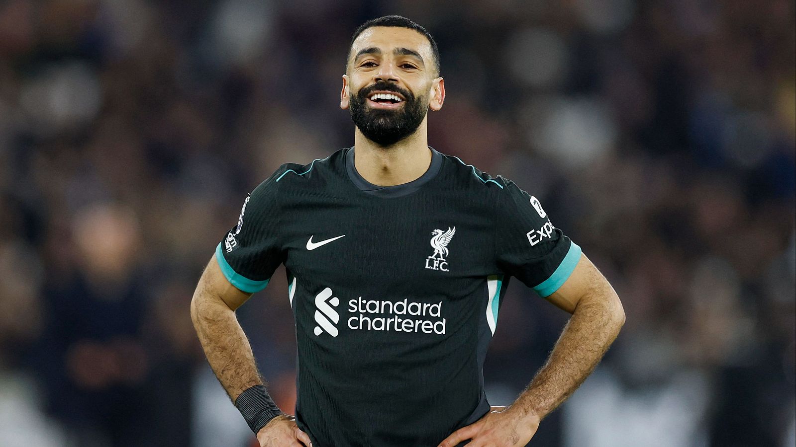 Mohamed Salah, con la camiseta del Liverpool, sonríe con las manos en las caderas.  Se aprecia el logo de Nike y el patrocinador en su uniforme.