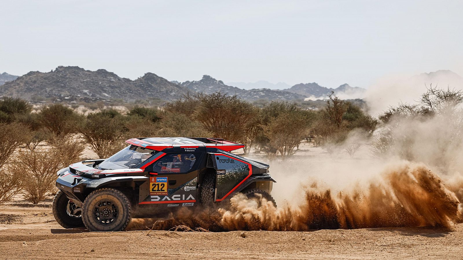 Coche de rally negro y rojo (nº 212) en carrera desértica, levantando polvo.  Patrocinadores visibles: Dacia y Motul.