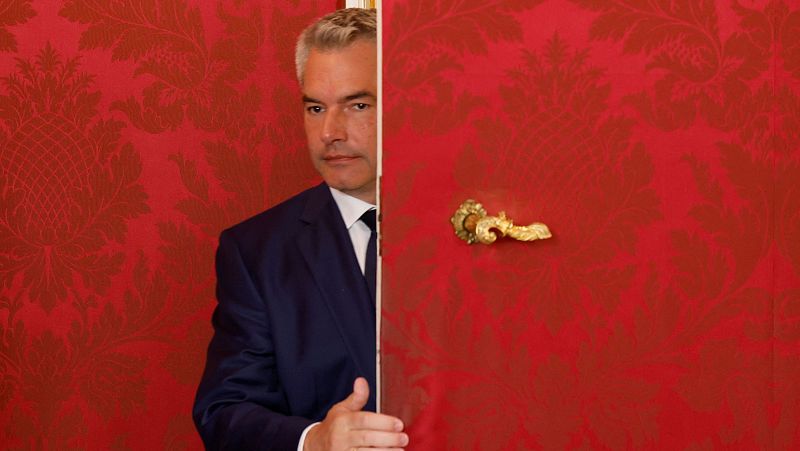 Excanciller austriaco, traje azul oscuro y corbata negra, asomándose por una puerta roja con relieve floral en la pared. Expresión seria.