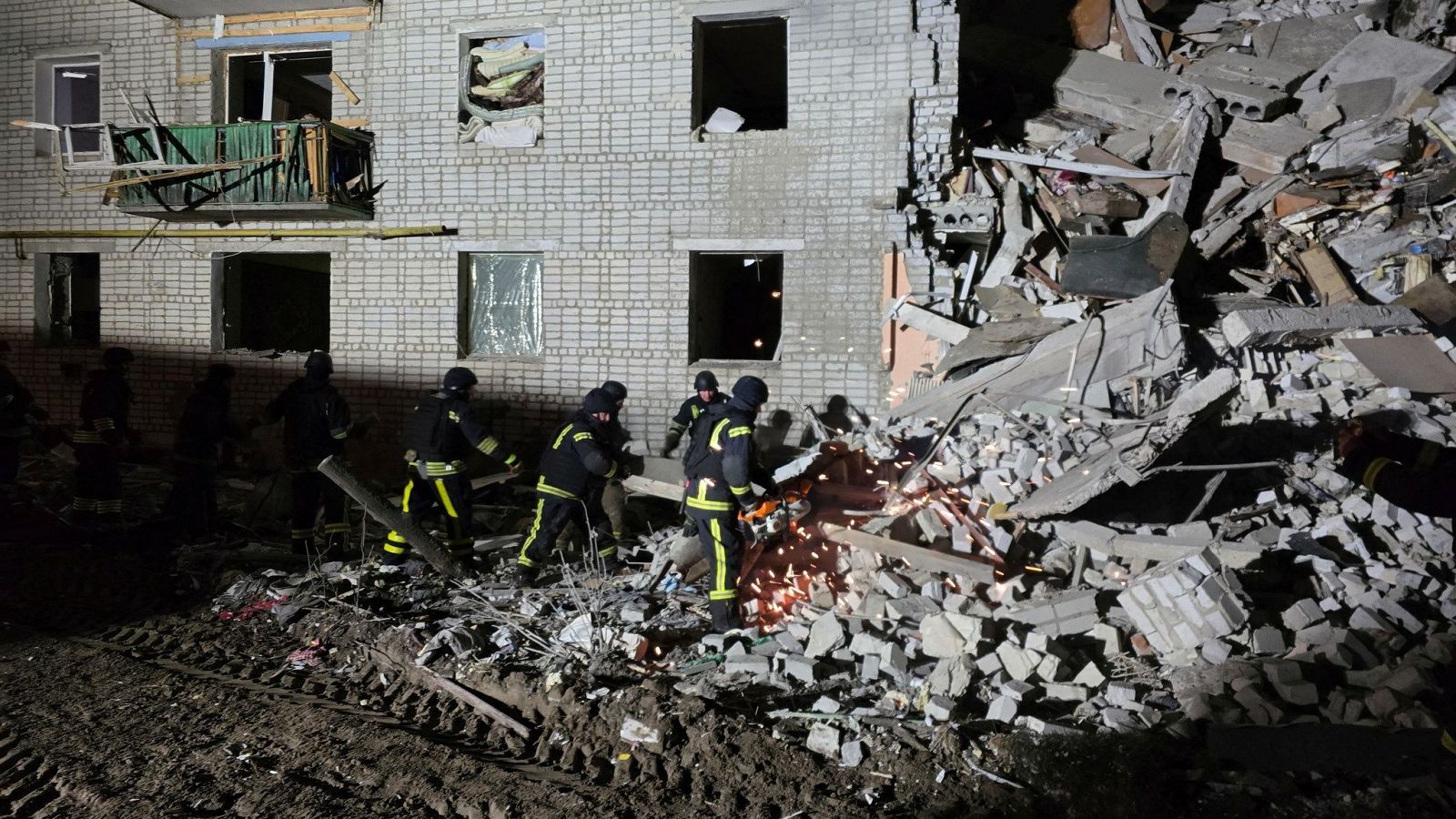 Bomberos trabajan entre escombros de un edificio dañado tras un bombardeo nocturno.  Se aprecian daños considerables en paredes y un balcón.