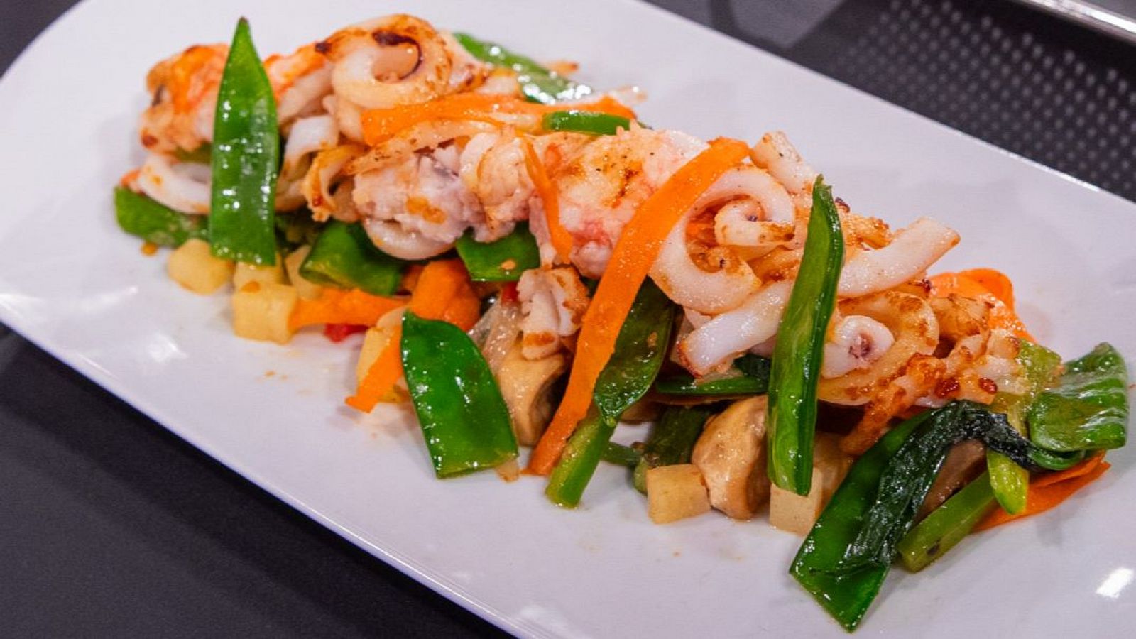 Ensalada de marisco y verduras en plato blanco rectangular.  Marisco salteado o a la plancha con verduras como zanahoria, guisantes y patata.