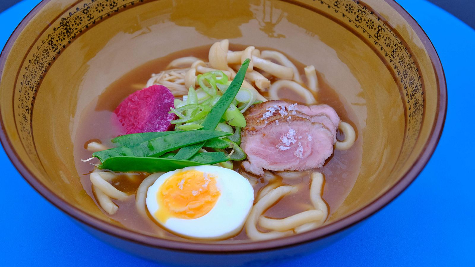 Ramen con fideos udon, cerdo ibérico, huevo, verduras y setas en un caldo marrón claro, servido en un cuenco.