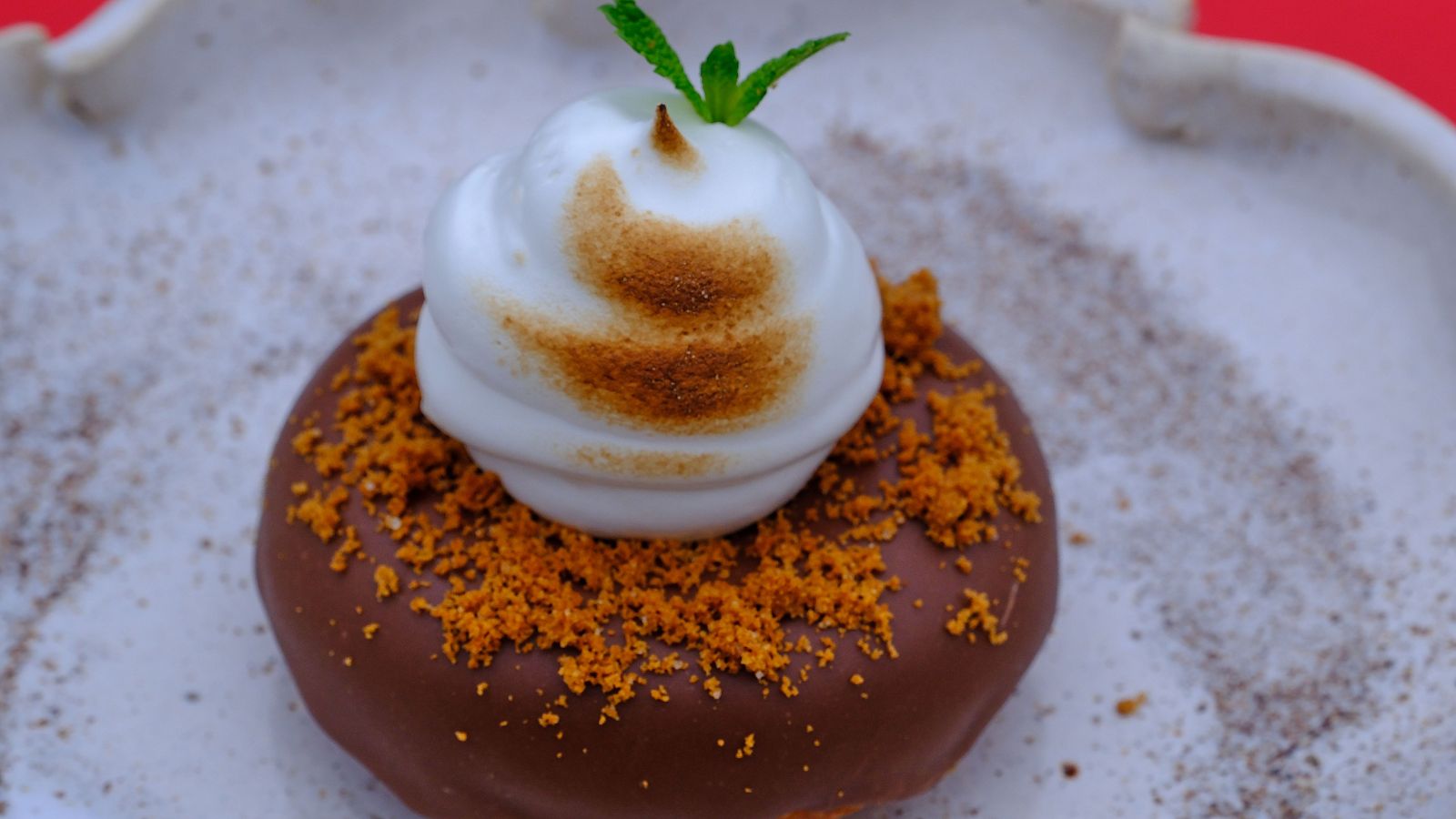 Donut con cobertura de chocolate, merengue blanco, menta y migas de galleta naranja.  Se encuentra en un plato blanco irregular sobre un fondo rojo.