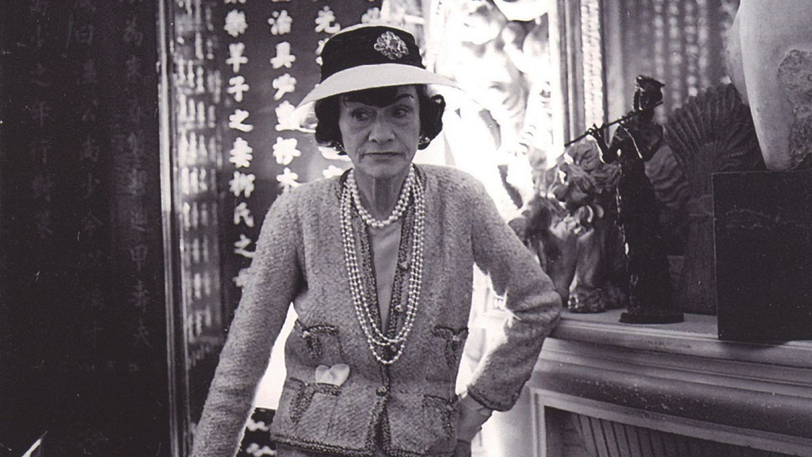 Coco Chanel en foto en blanco y negro, con chaqueta de tweed, collares de perlas y sombrero; fondo con biombo oriental y repisa con decoración.