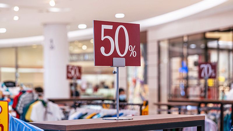 Cartel rojo con texto blanco anuncia un 50% de descuento en rebajas. Se observa en una mesa de madera oscura, con otros carteles de rebajas en el fondo.