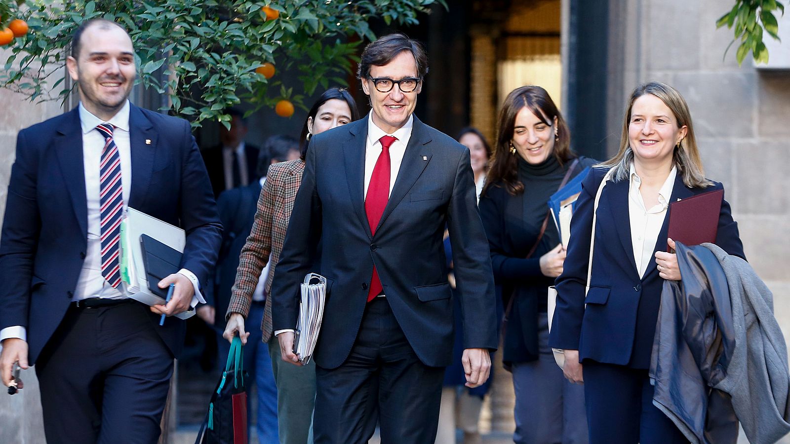 El president del Govern, rodeado de consejeros, aparece en la entrada de un edificio.  Visten formalmente; él lleva traje oscuro y corbata roja.