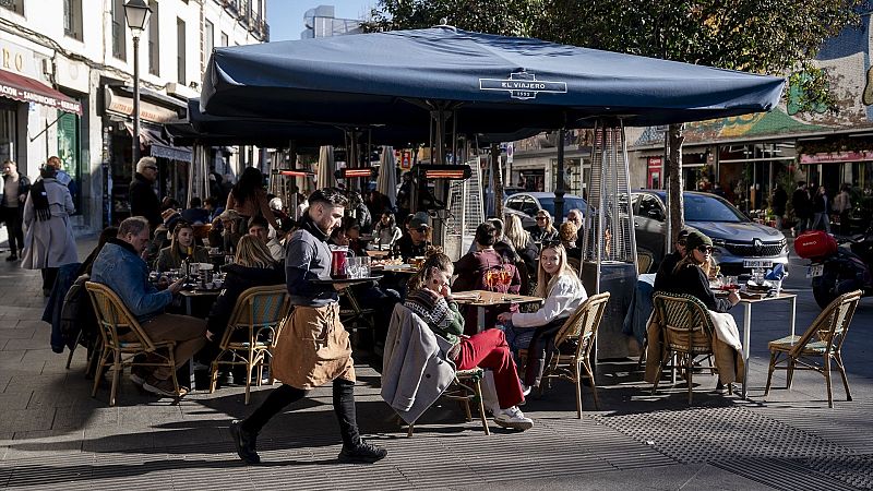 Terraza de restaurante llena de gente, con mesas, sombrillas y un camarero sirviendo. Ambiente soleado y animado.