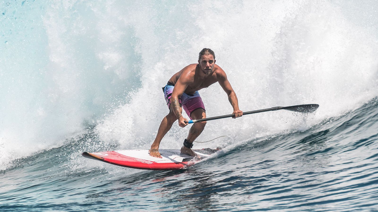 Hombre en paddle surf sobre tabla roja y blanca, con remo, cabalgando una ola. Lleva pantalones cortos estampados y el pelo recogido.