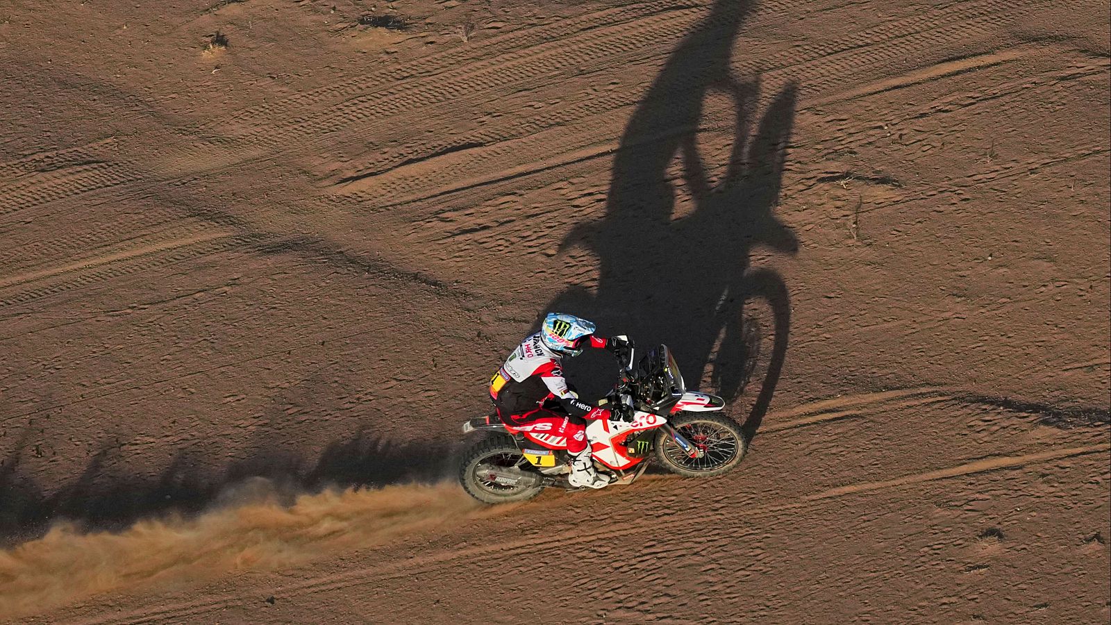 Motociclista en carrera de motocross desértica, inclinado en curva, levantando polvo con su moto roja y blanca.  Se ven marcas de neumáticos en el suelo.