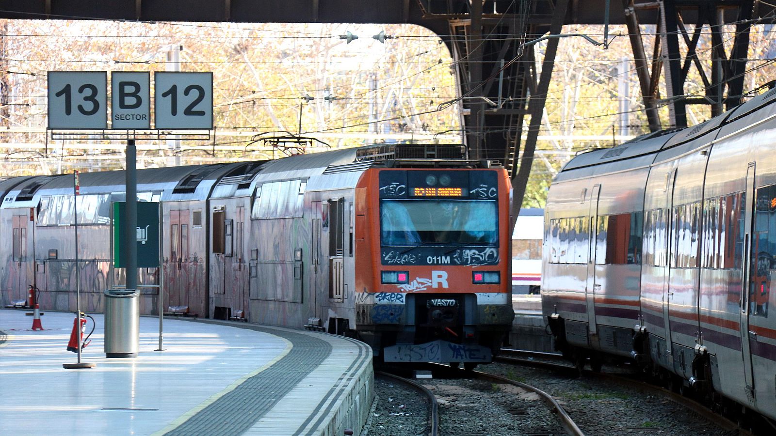 Tren Rodalies (011M R) saliendo de estación, con grafitis.  Se ve letrero '13 B 12 SECTOR' en andén.