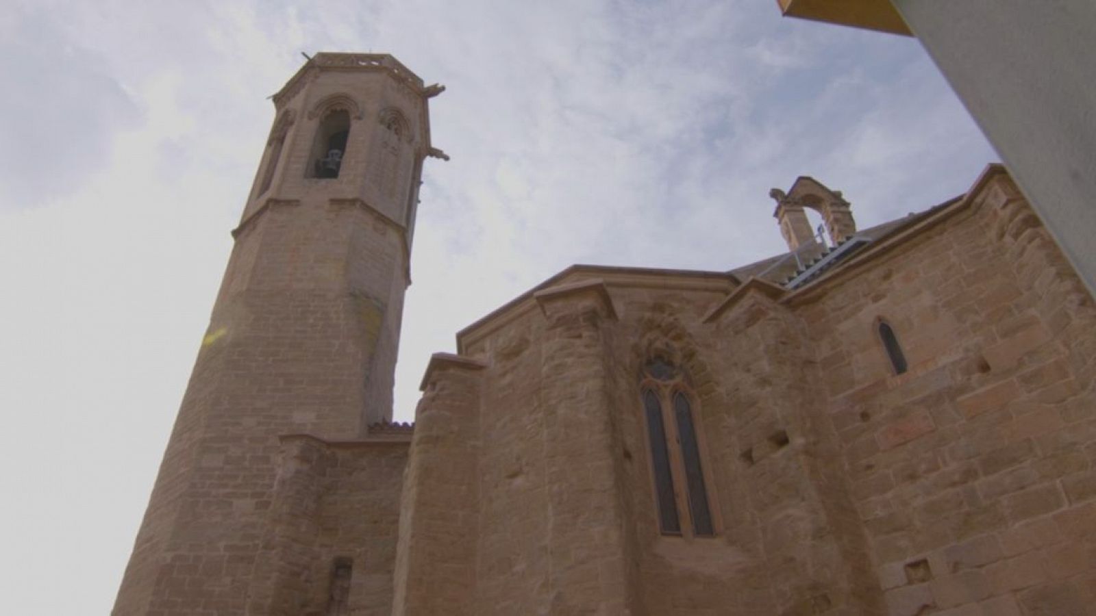 Vista de la torre octogonal de una iglesia, construida con piedra marrón clara, con detalles como aberturas superiores y posibles gárgolas.  Se observa también una sección adyacente con una ventana alta.