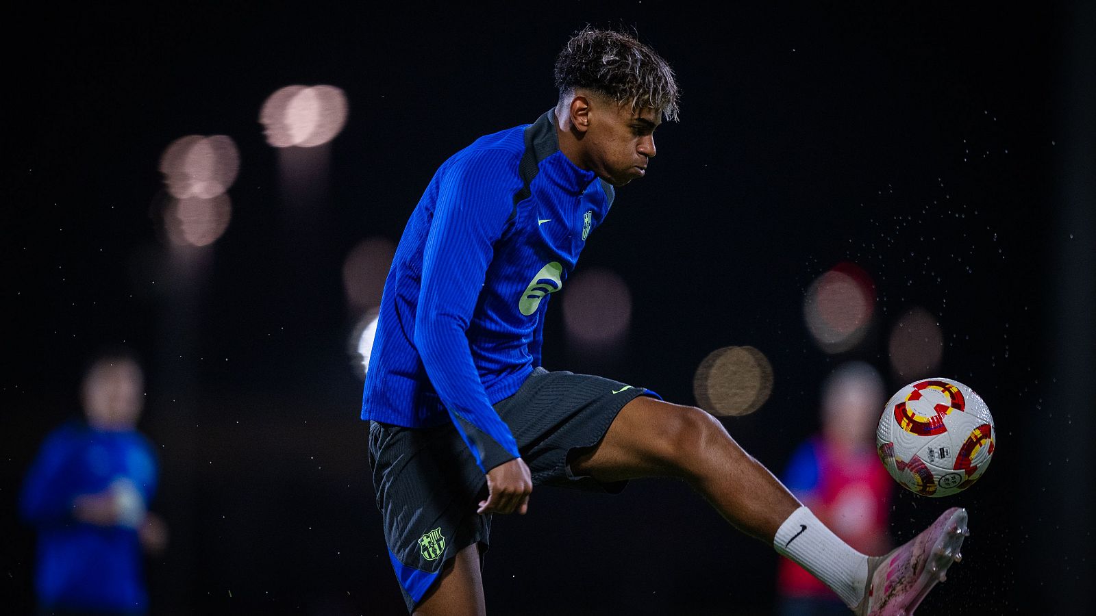 Lamine Yamal, con la equipación del Barça (azulgrana con detalles verdes y pantalón negro), controla un balón en un campo de fútbol iluminado artificialmente.  Se aprecian otros jugadores en el fondo.