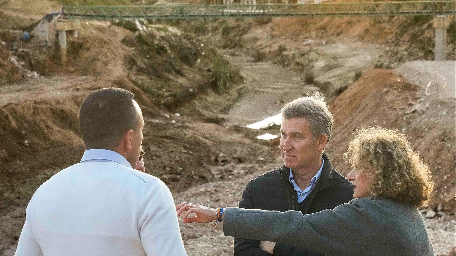 Visita de Alberto Núñez Feijóo a una zona afectada por la DANA en Valencia.  Terreno dañado, puente al fondo, acompañado de dos personas.