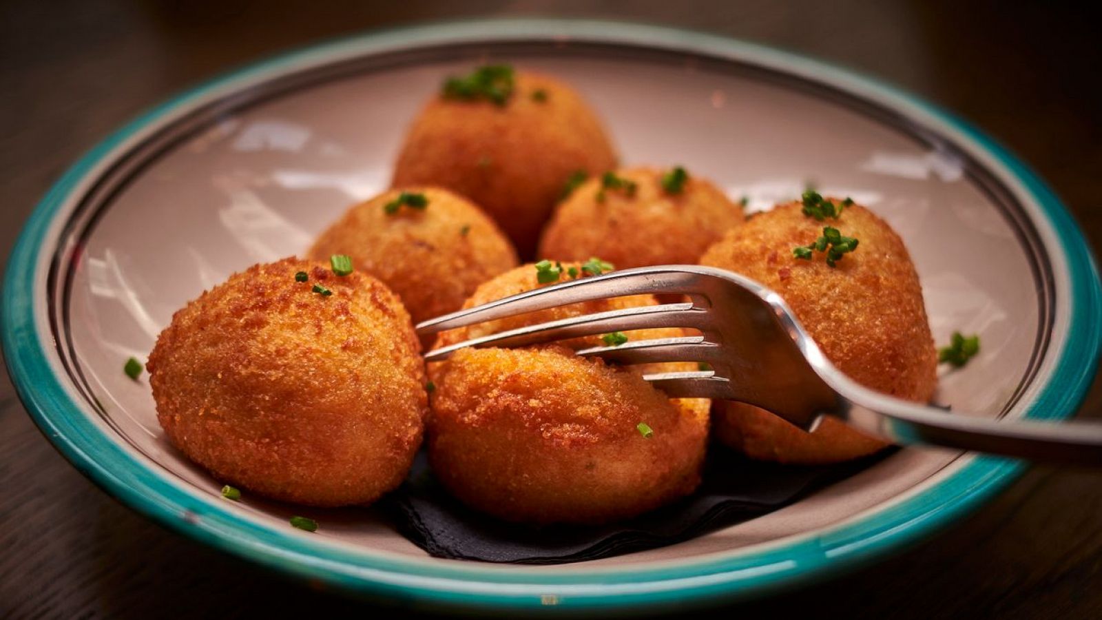 Croquetas doradas y crujientes, espolvoreadas con cebollino, en plato cerámico turquesa y beige.  A punto de ser comidas.