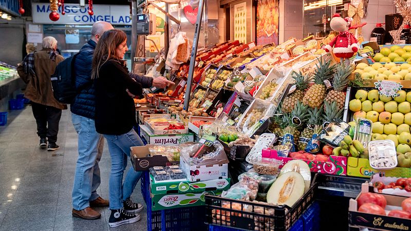 Mercado con frutas, verduras y mariscos congelados. Pareja observa productos frescos;  una caja dice "El auténtico".