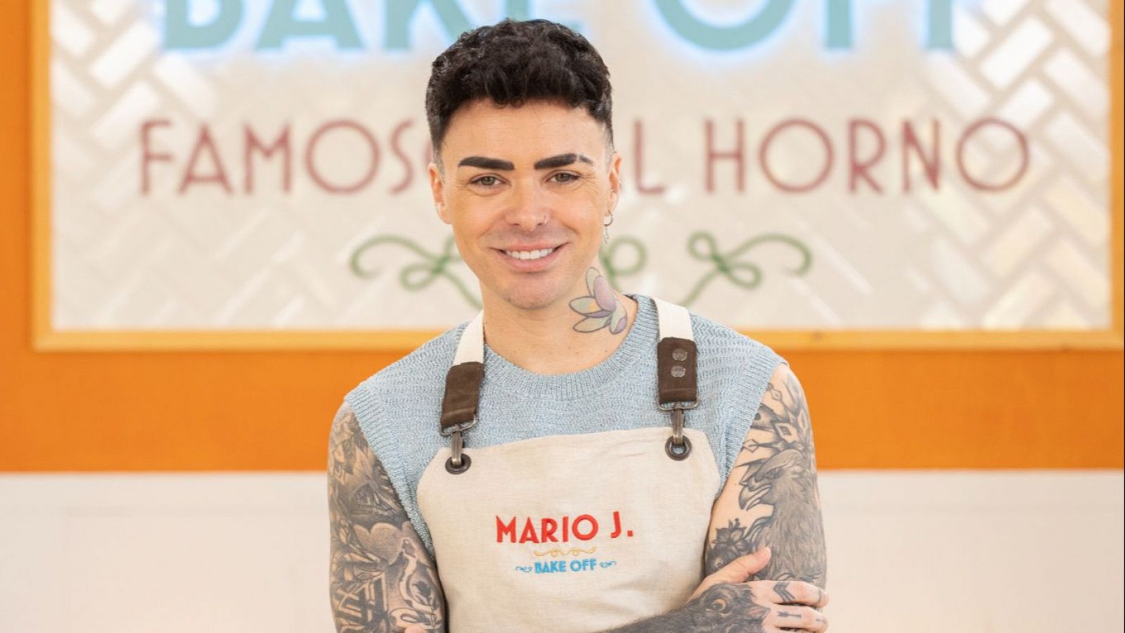 Mario Jefferson, de 'Bake Off: Famosos al horno', sonríe con los brazos cruzados, luciendo camiseta azul, delantal beige bordado y tatuajes. Fondo con letrero del programa.