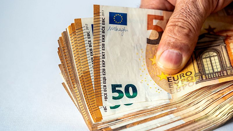 Mano con arrugas sujetando un fajo de billetes de 50 euros (año 2017 visible), mostrando marcas táctiles. Ilustración de un incremento económico.